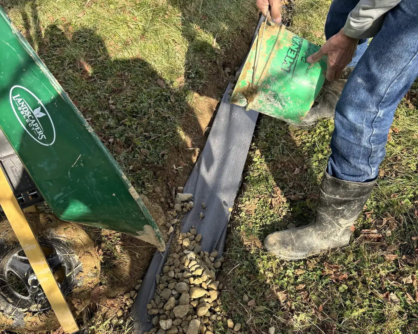 Corrugated drainage pipe and filter fabric being set into a hand-dug trench beside a loaded wheelbarrow
