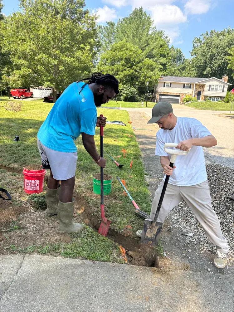 Two Hoosier Water Away crew members opening a drainage trench with shovels beside a driveway