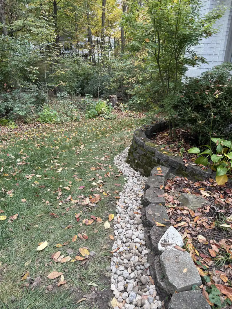 Finished trench drain of river rock curving along a stone retaining wall beside the lawn