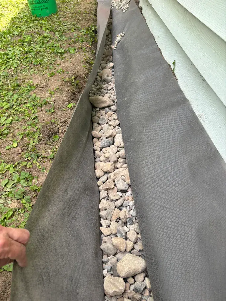 Hand holding back filter fabric along a stone-filled trench next to a house