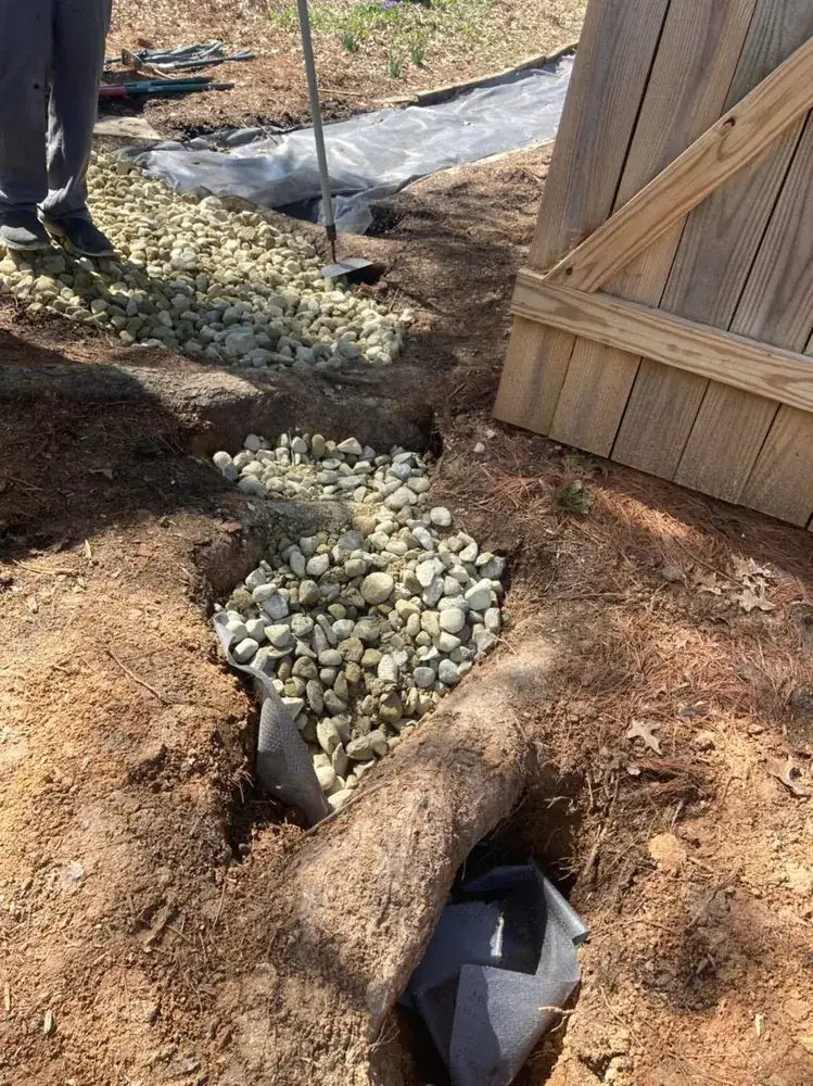 Trench dug by hand along a wood fence, with stone laid in around a tree root that was left in place