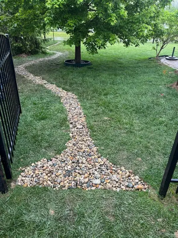 Finished river rock drainage channel curving across a green lawn, through an iron gate and around a tree ring