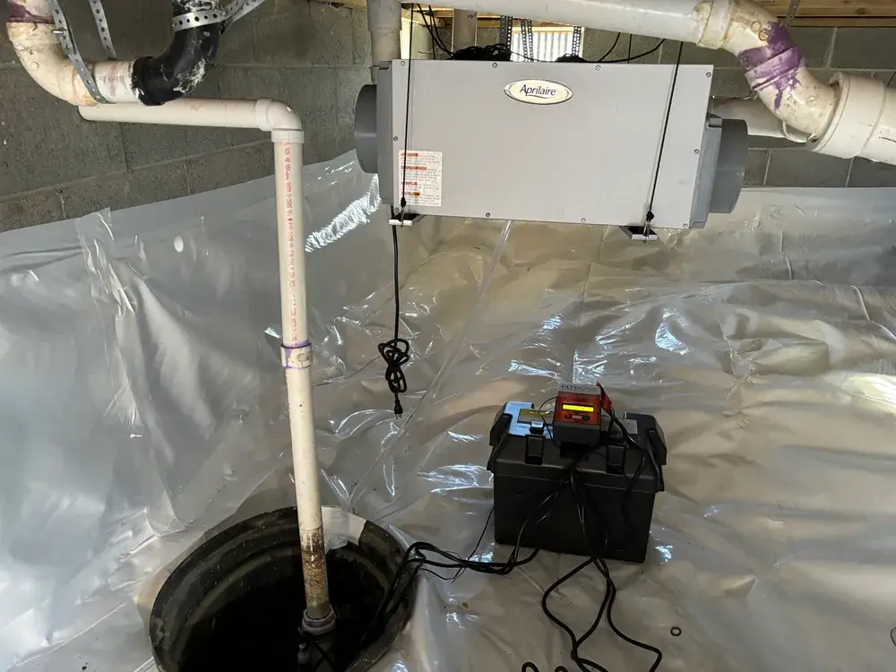 Sump pit with discharge pipe and a battery backup box on white vapor barrier, with a wall-mounted dehumidifier above