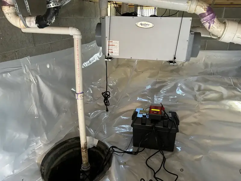 Sump pit with discharge pipe and a battery backup box on white vapor barrier, with a wall-mounted dehumidifier above