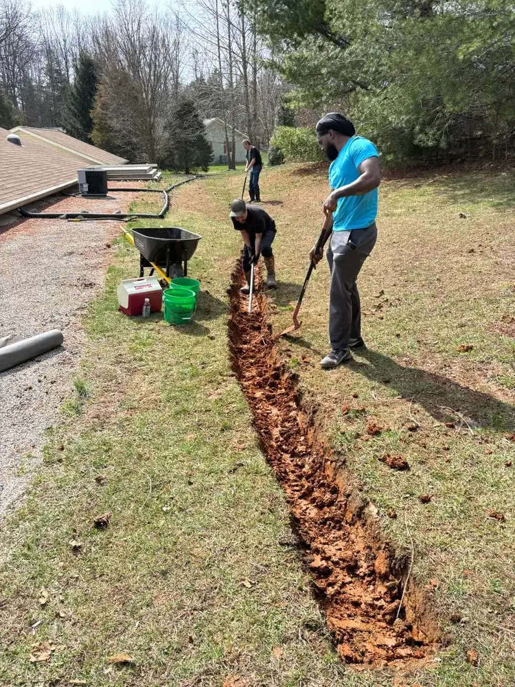 Crew digging a drainage trench by hand across a sloped lawn beside a gravel driveway